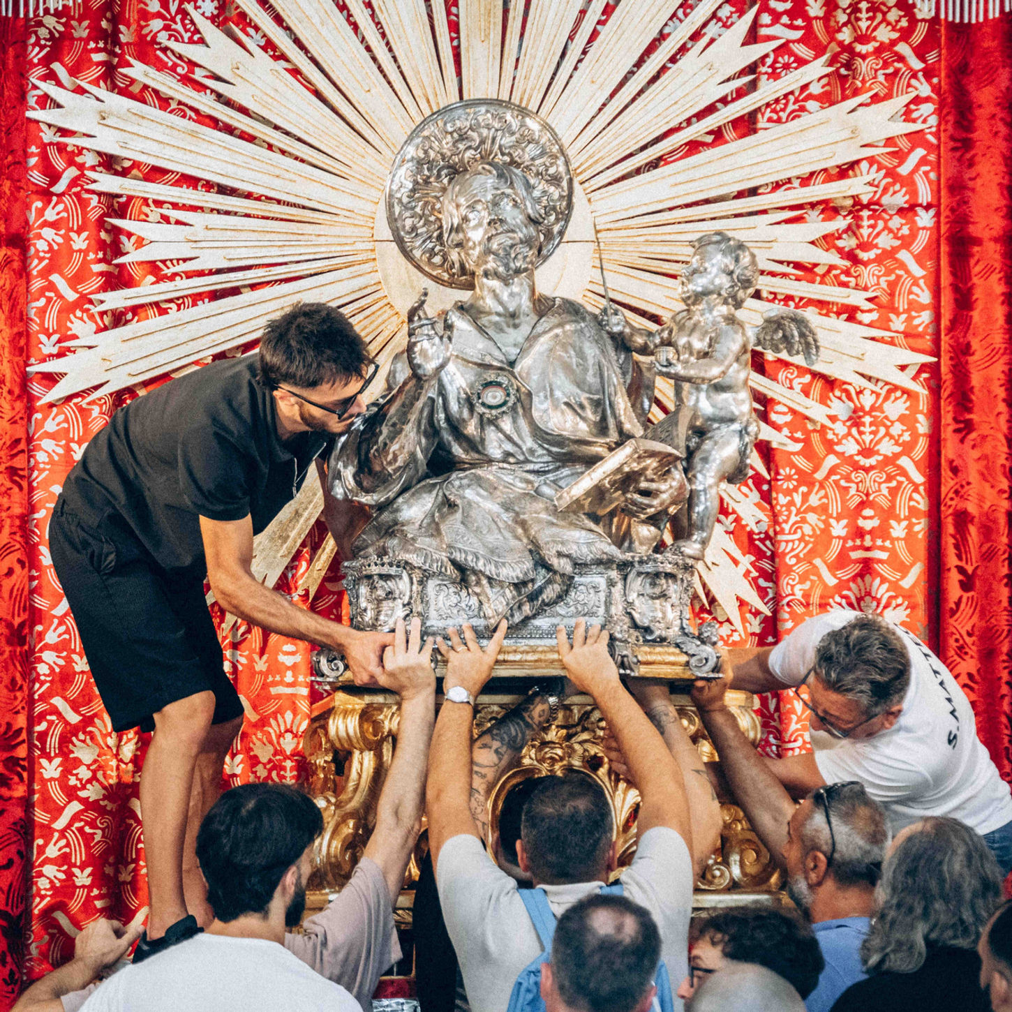 A group of people carefully lifting and carrying a silver religious statue in front of a red draped backdrop.