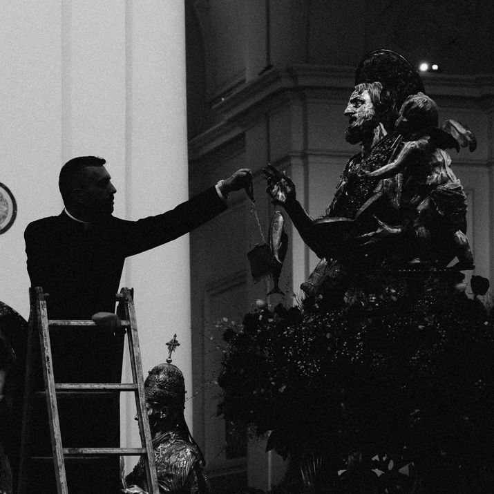 Black and white photograph of a man on a stepladder adjusting a large religious statue inside a church.