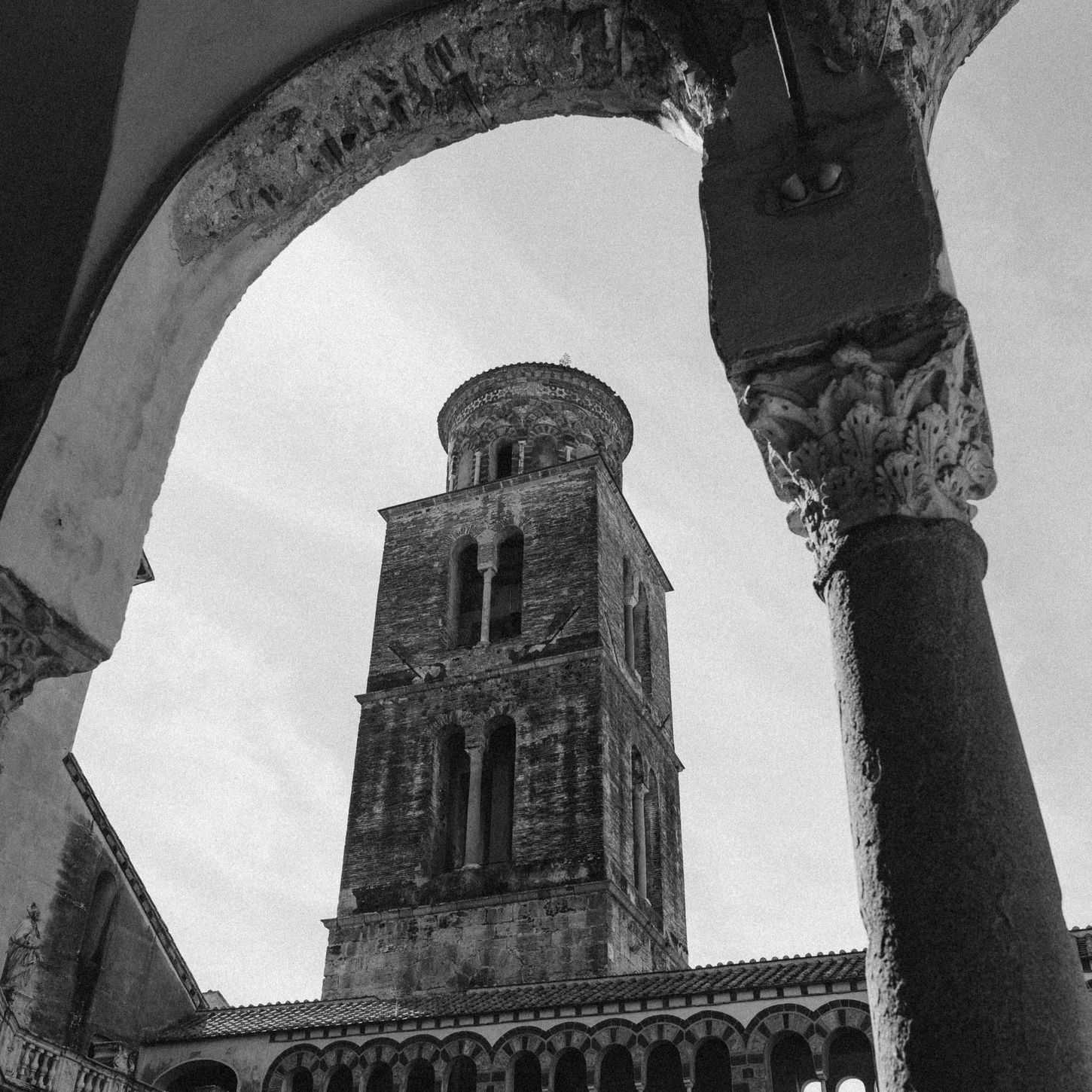 Black and white low-angle photograph of an ancient stone tower framed by arched corridors and historic columns.