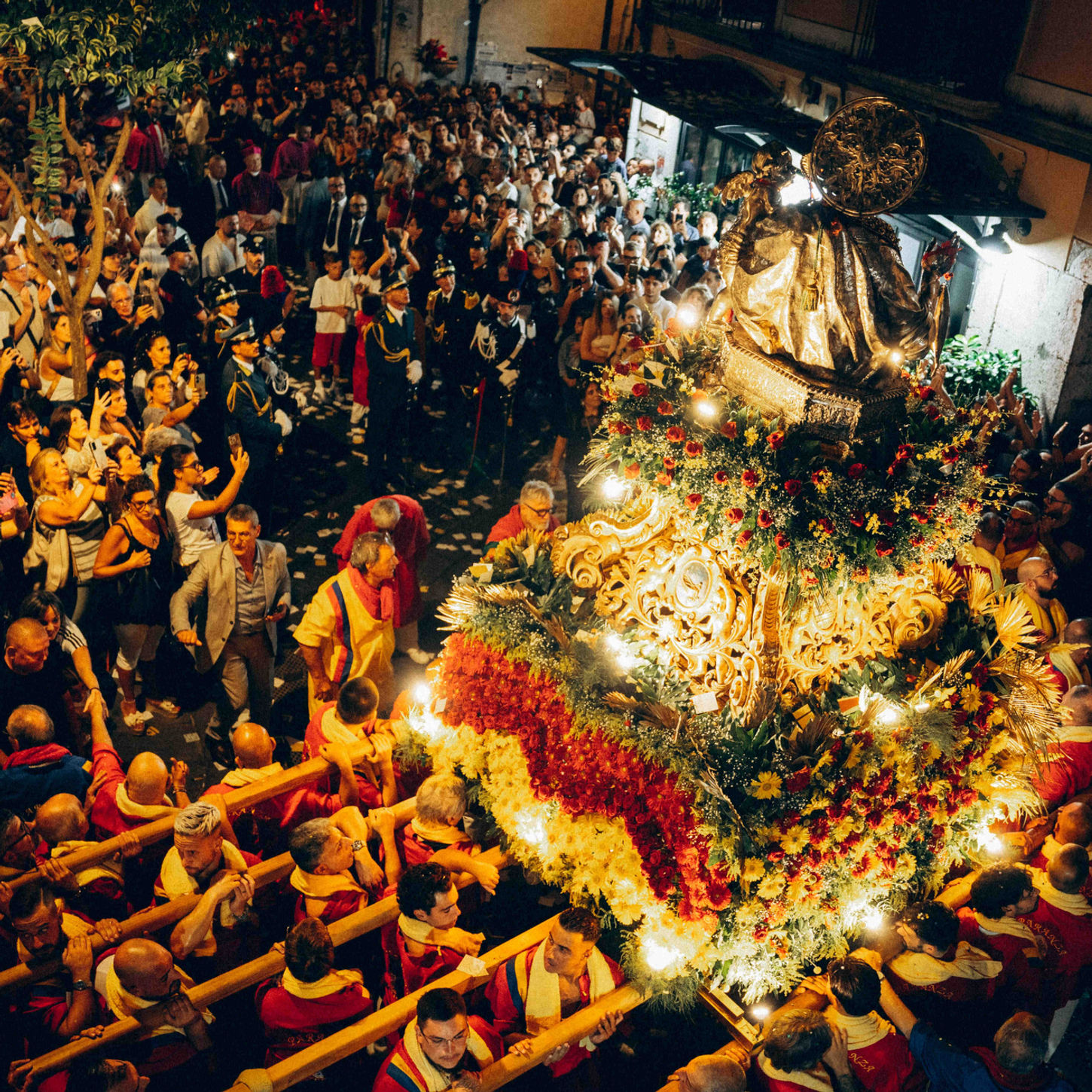High-angle shot of a crowded street filled with people gathered around an illuminated religious float decorated with flowers at night.