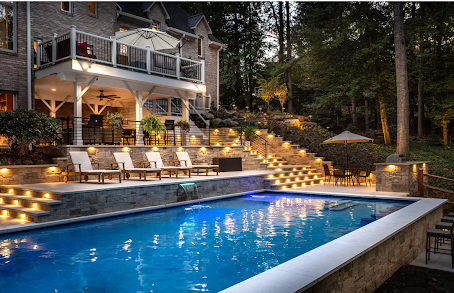 Backyard pool area with illuminated stone steps, patio seating, and raised deck by NOVA Landscape Construction