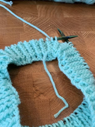 Knitted aqua hat body on circular needles, early shaping stage with yarn tail visible