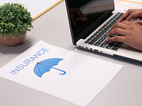 Close-up of a person typing on a laptop beside a document labeled “Insurance” with an umbrella icon, representing coverage and financial protection, on a clean office desk with a small plant.