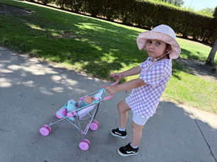 Olive Mae looking at the camera wearing a sun hat and checkered shirt pushing a baby doll stroller.