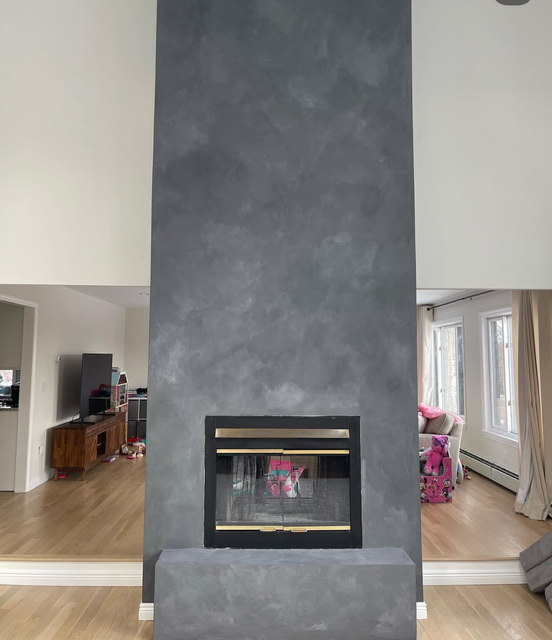 Modern living room with a tall gray lime wash plaster fireplace wall, black-framed hearth, and pink kids’ toys by bright windows.