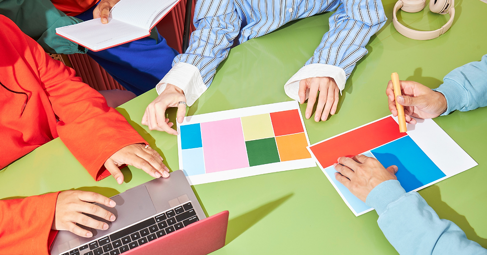 People at a table collaborate creatively, surrounded by colourful paper, a laptop, and headphones. Bright, focused atmosphere with vibrant tones.
