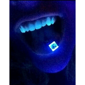 Primer plano de una boca abierta con dientes iluminados en azul; sobre la lengua se ve un pequeño cuadrado de papel tipo blotter con un diseño.