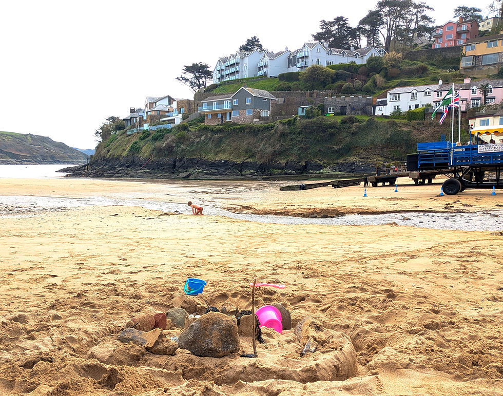 Sand castle, bucket and spade on South Sands, Salcombe. Sea tractor in middle ground, cliffs with colourful houses in background. View of estuary.