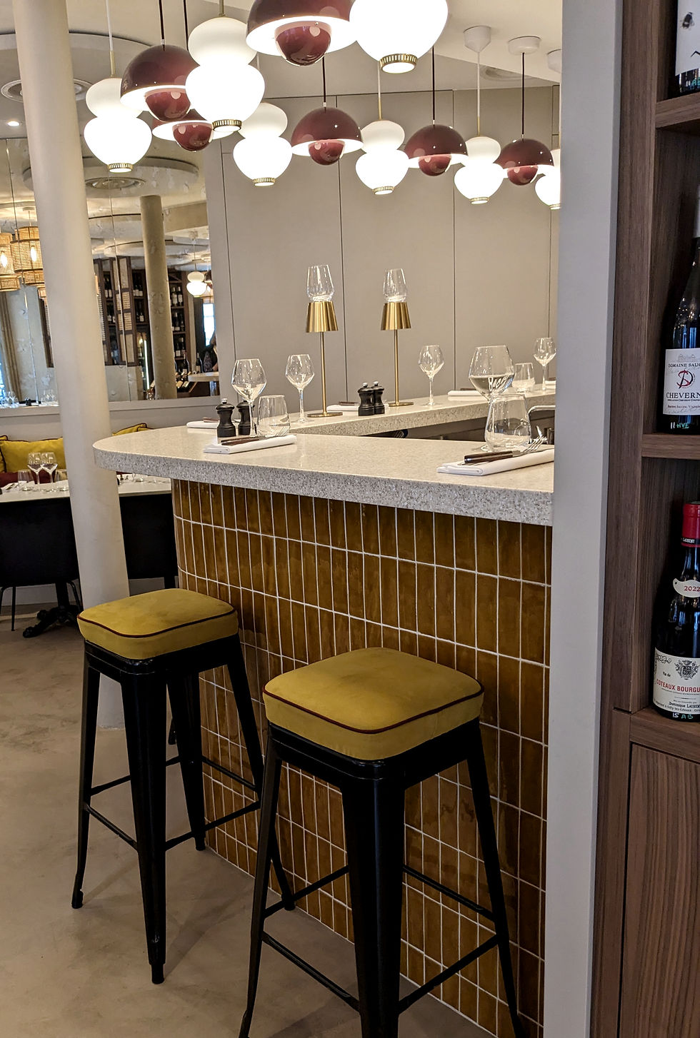 A chic Parisian bar counter at Chonbou restaurant featuring mustard yellow tiles, matching velvet bar stools, and modern globe pendant lights."