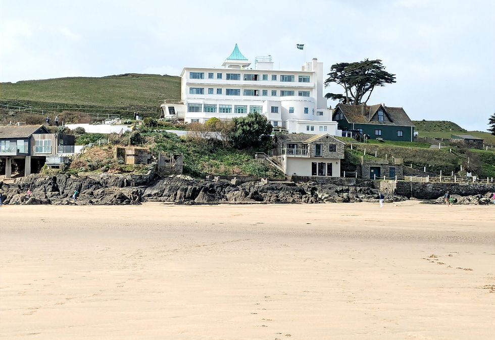 A view of 3 storey white hotel in the style of Art Deco sitting atop the stone ridge of Burgh Island. Foreground is a wide expanse of sandy beach.