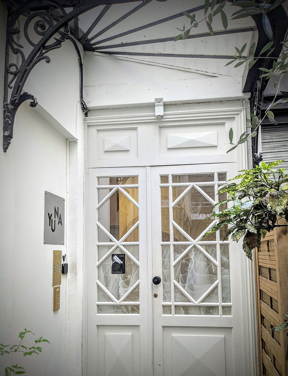 Chic white front door with diamond shaped beading in two glass panels. The doorway has a sign on it saying Yuna Blanche Apartments. There is a gold bell, black wrought iron detailing and green foliage.