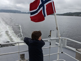 An autistic boy stands at the back of a boat on a fjord tour leaving from Bergen. He looks at the water and mountains. The Norwegian flag flies above him.