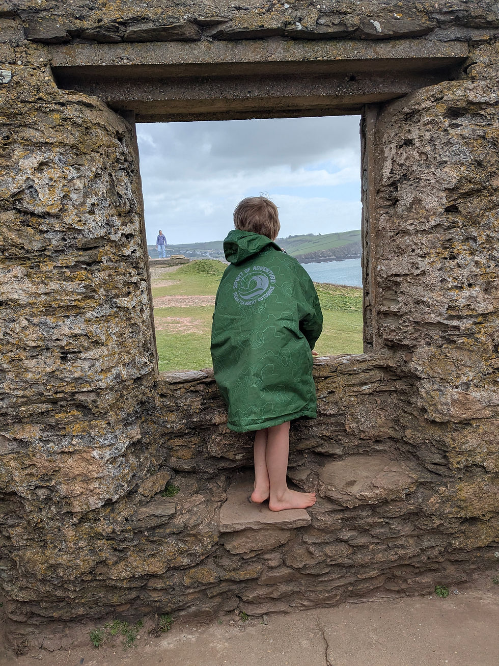 An autistic child wearing a green dry robe looks out of a stone window cavity to the view of sea and cliffs. Standing on thick stone ledge inside ruined chapel at the top of Burgh Island.