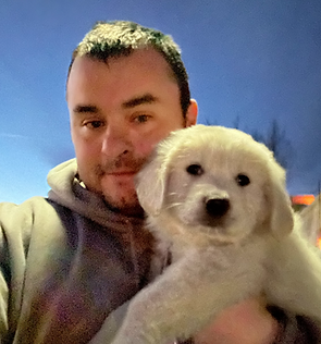 Man in grey hoodie holding white Golden Retriever puppy outdoors.