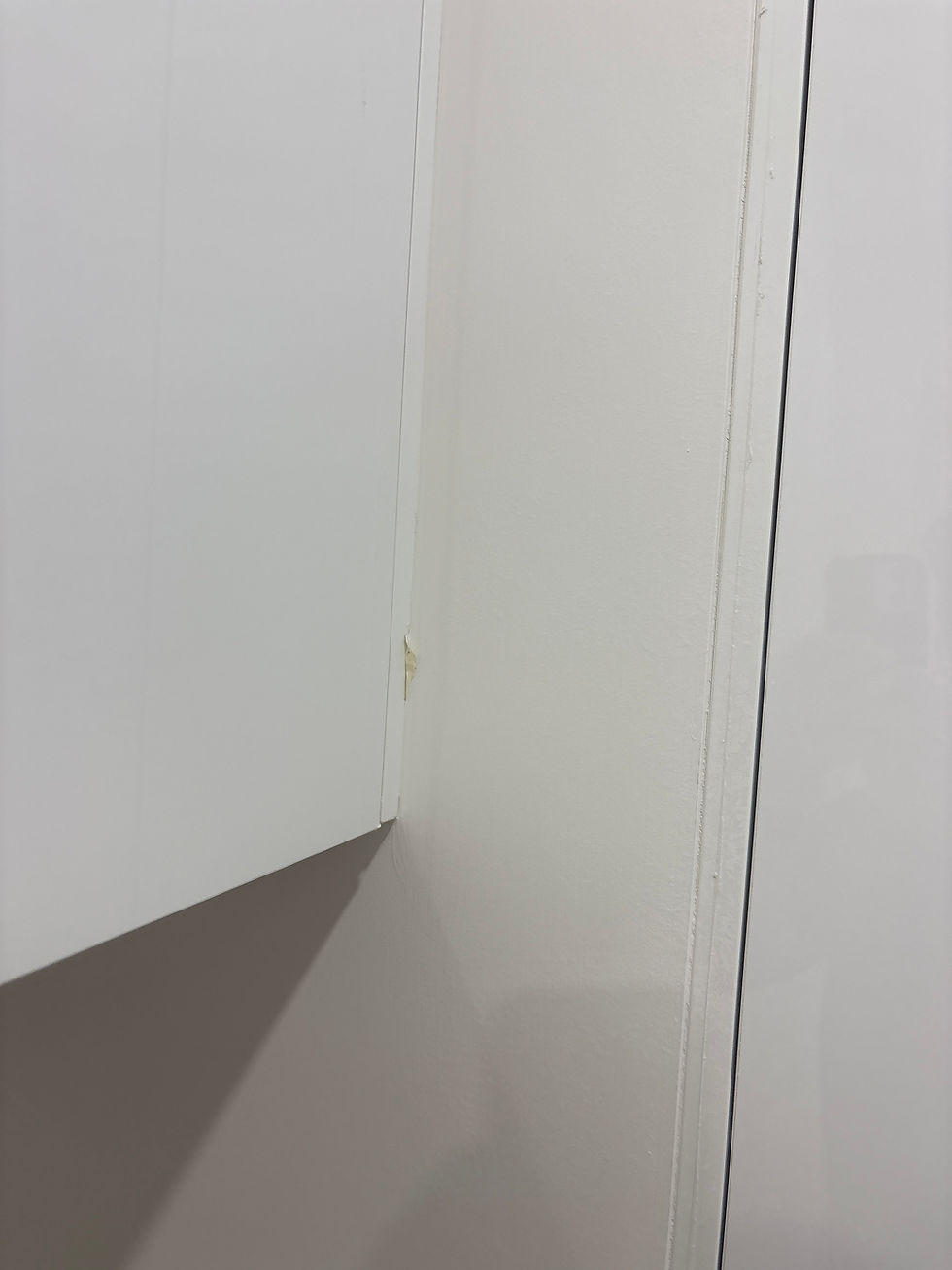 Close-up of a white cabinet door corner against a white wall, showing a small chip or damage. The setting appears minimal and clean.