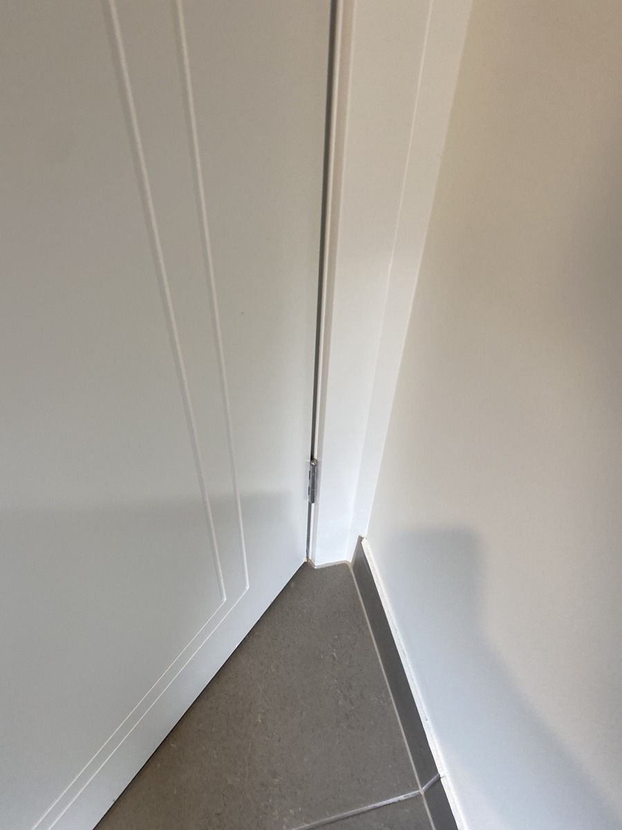 White door slightly ajar in a corner, casting a shadow on light gray tiled floor. Walls and door have simple linear patterns.
