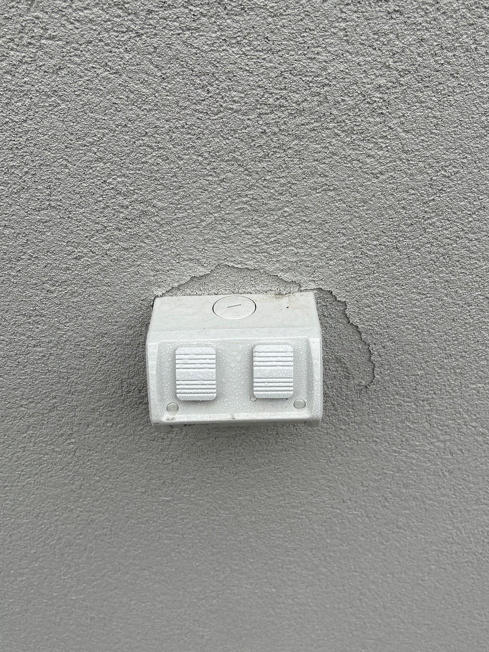 Gray wall with a textured surface featuring a white, weatherproof outlet cover. Water droplets visible, indicating recent rain.