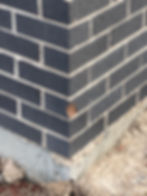 Corner of a building with dark gray bricks and light mortar, one chipped. Concrete base visible, surrounded by dirt and debris.