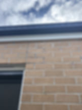 Brick wall with a dark window in the lower left, beneath a gutter. Blue sky and clouds are visible above, creating a calm scene.