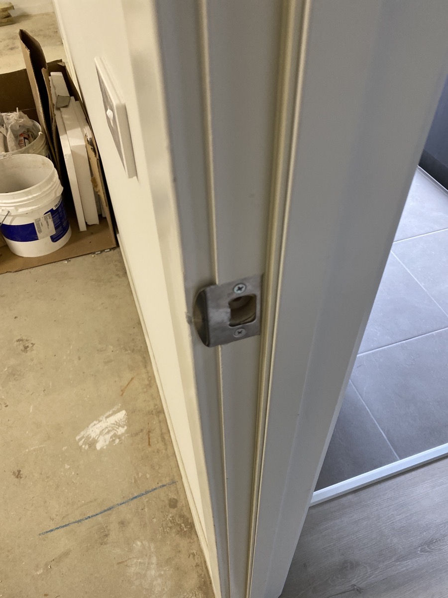 Door close-up with latch and light switch on a white wall. Buckets and tools are seen on the concrete floor, with a tiled room beyond.