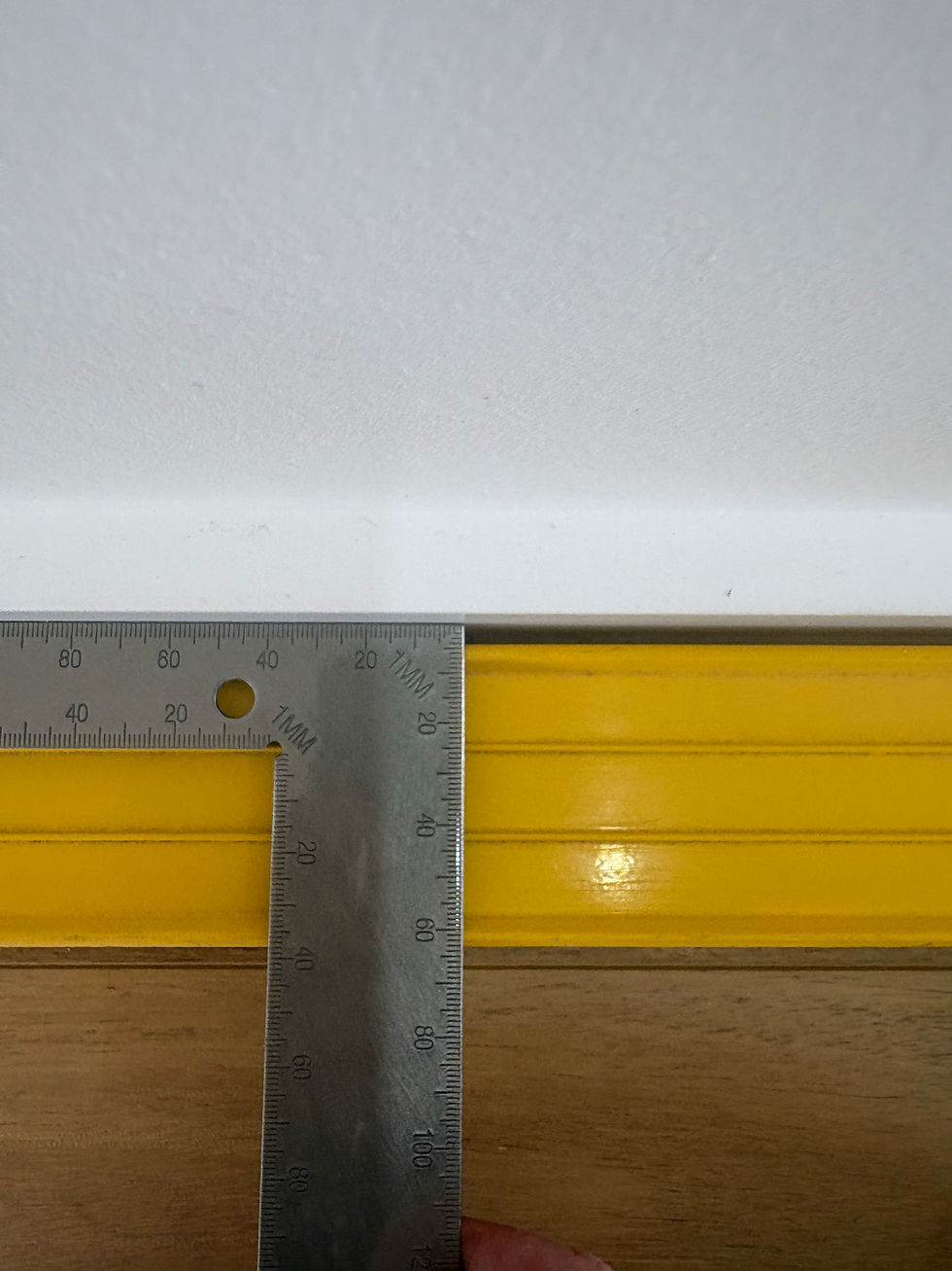 Metal ruler measuring a white wall and yellow trim. The background is wooden, and ruler markings are visible. Calm, precise setting.