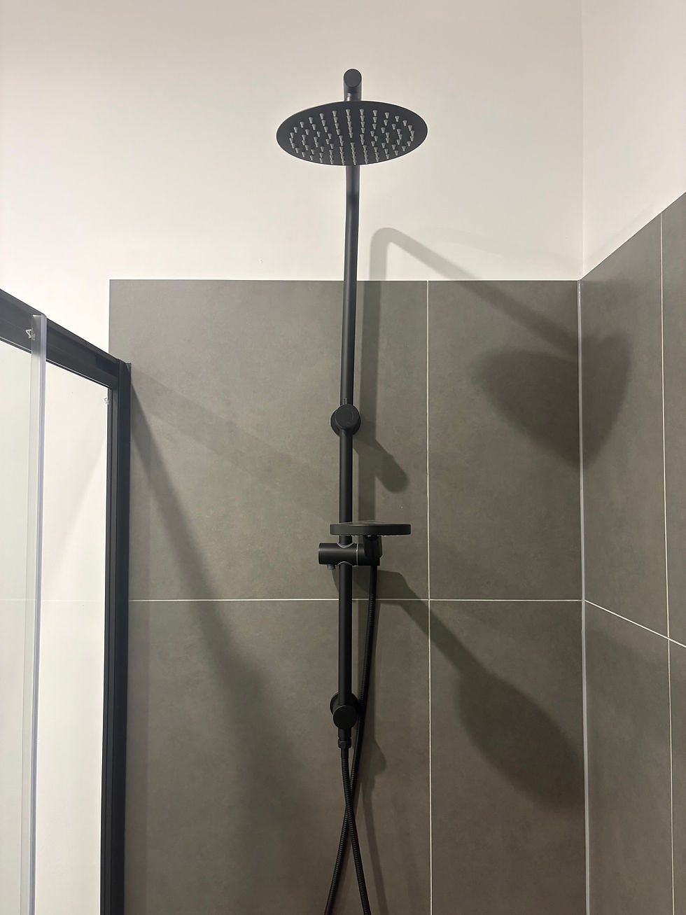 Black rain showerhead and control panel against gray tiles in a modern bathroom. Glass shower door partially visible. Minimalist design.
