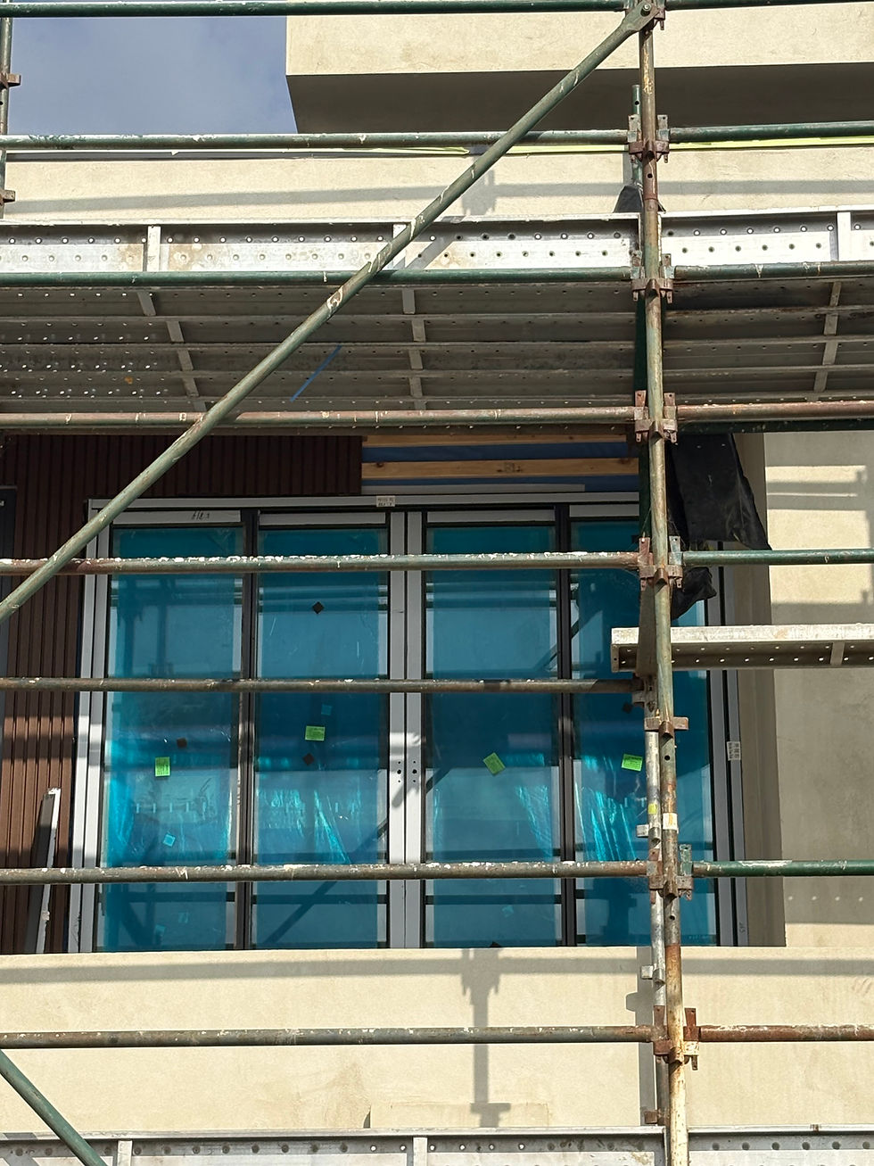Scaffolding in front of building facade with teal-covered windows and beige walls. Clear sky above, construction setting visible.