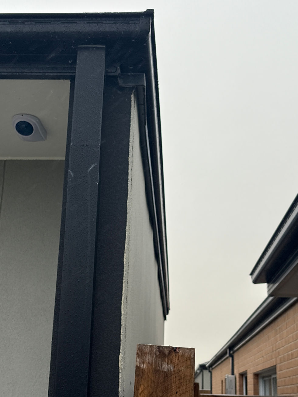 Building corner with black trim, surveillance camera, and adjacent brick wall under overcast sky. Wooden post visible in foreground.