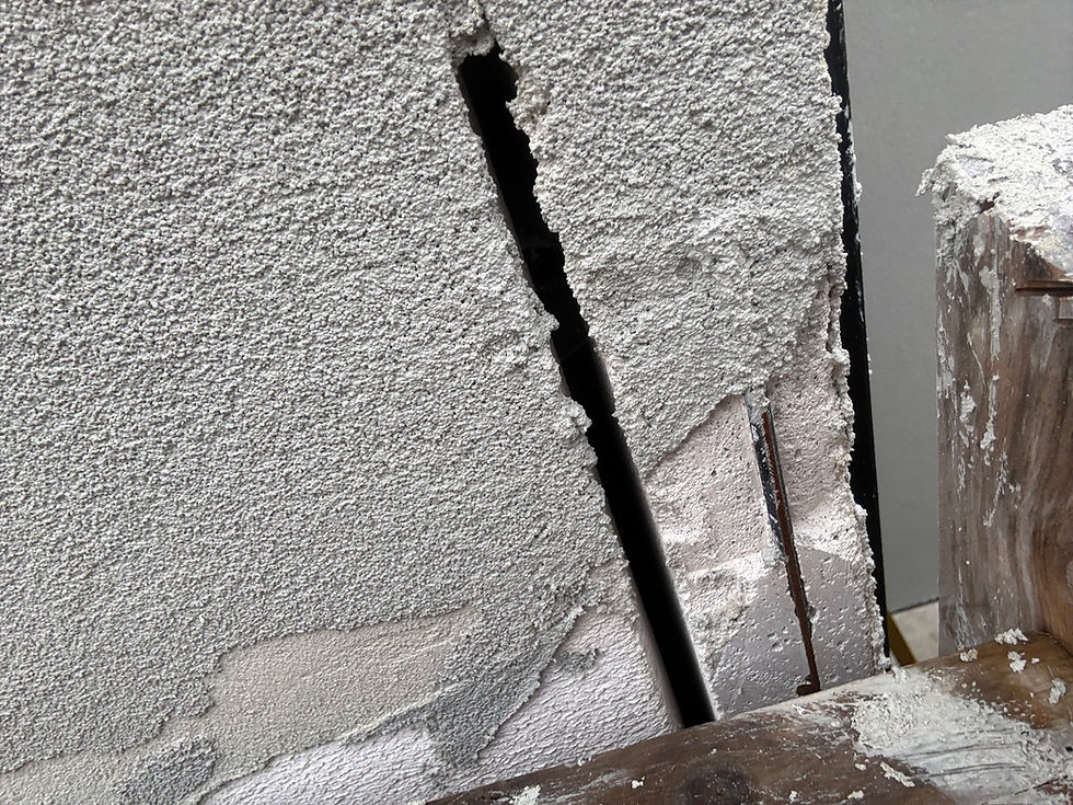 Close-up of a wall with a large crack, showing exposed reinforcement bars. The surface appears rough and weathered, with visible debris.