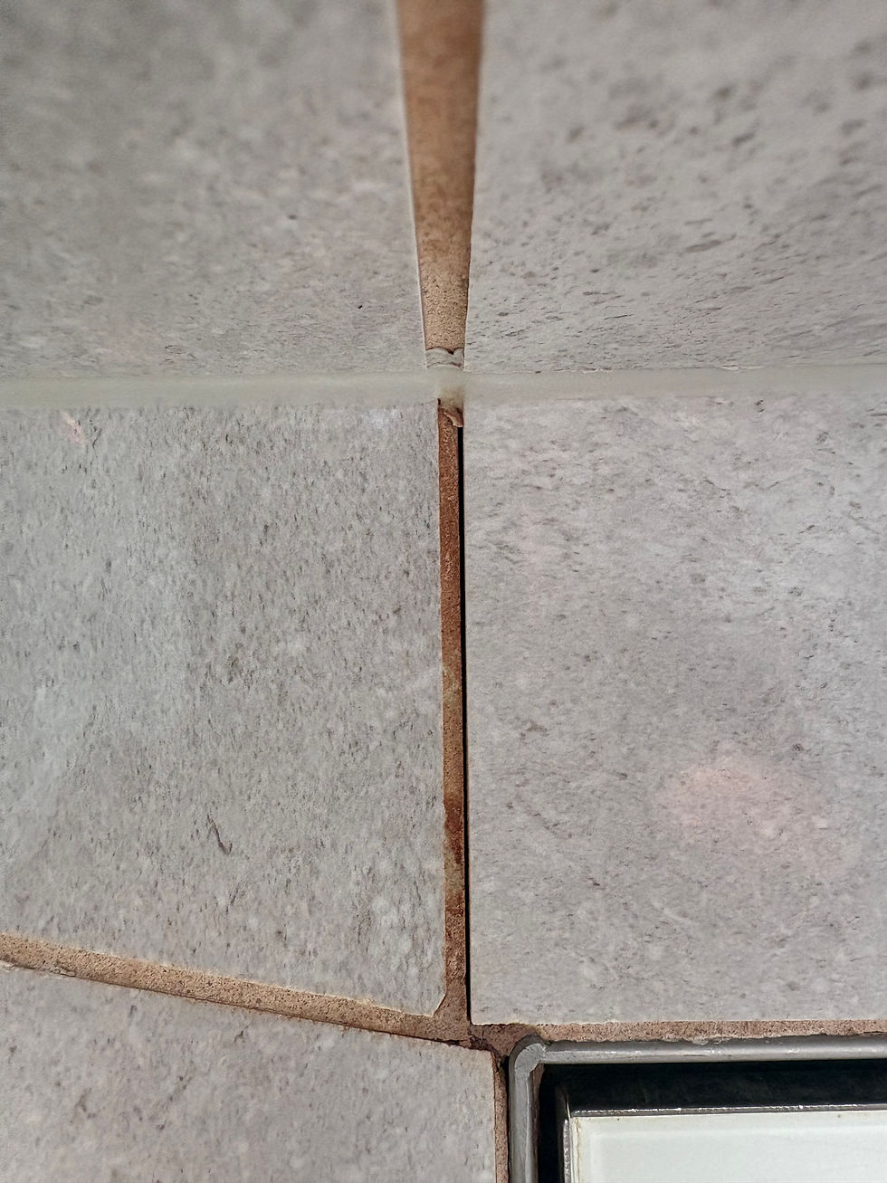 Close-up of gray, textured tiles with visible grout lines. A white object is partially visible in the bottom right corner.