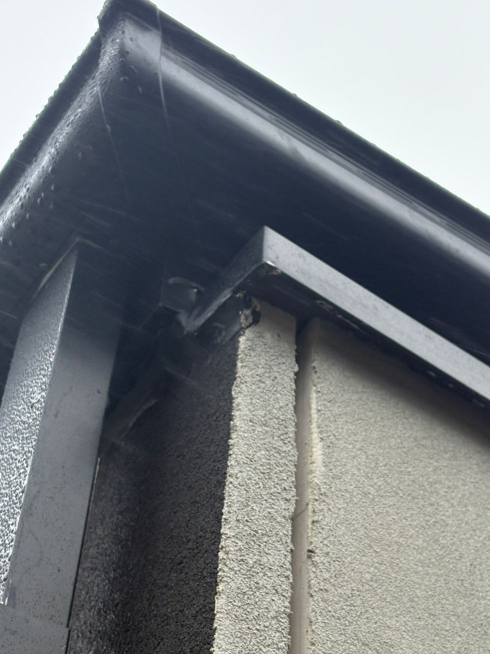 Black rain gutter on a building corner with textured gray and black wall surfaces. Overcast sky in the background.