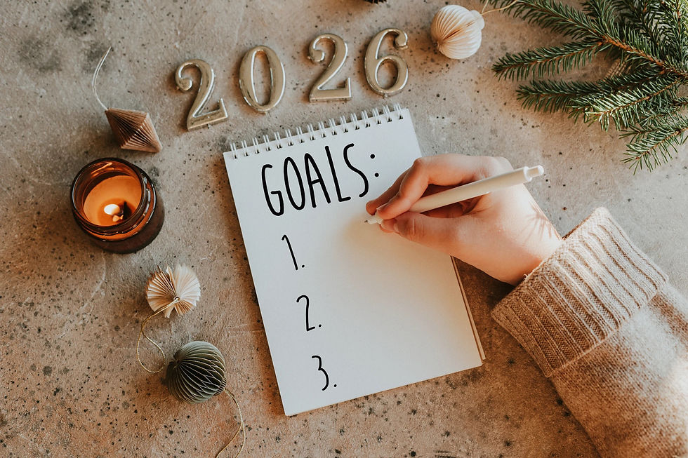 Hand writing on a "GOALS" notepad with "2026" in silver, surrounded by a candle, ornaments, and pine branch on a stone surface. Cozy mood.