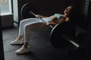 A woman wearing a white athletic set and white shoes completing a barbell hip thrust.