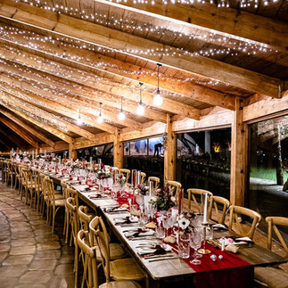 Longue table de réception élégamment dressée sous une charpente en bois, éclairée par des guirlandes lumineuses, avec vaisselle raffinée et décorations florales, dans une ambiance chaleureuse de soirée.