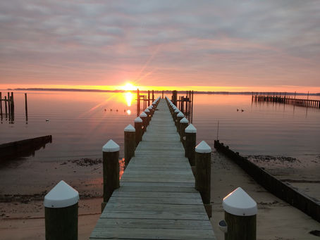 Sunrise over the bay with a wooden dock extending toward the horizon, the rising sun glowing just left of the dock and reflecting across the calm water.