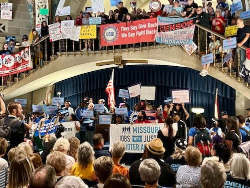 Missourians gather inside the Capitol, holding signs and banners advocating for workers' rights and social justice. The banners prominently display messages supporting unions and fighting back against censorship, with participants actively engaging in the demonstration.
