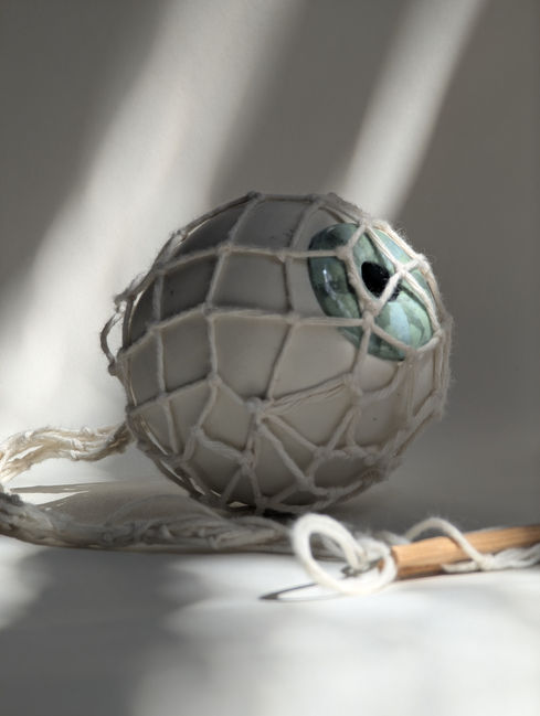 An oversized, simplified porcelain eye with a blue-grey striped iris, nested inside a hand-knotted white twine net bag. A length of twine extends from the back of the form like an optical nerve, snaking across a white backdrop to a handmade wooden net shuttle.
