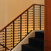 Modern staircase in a remodeled Sedona home featuring a warm natural wood handrail, custom geometric dark metal balusters, and dark treads against warm-toned walls.