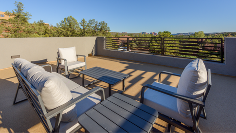 Modern outdoor sky deck of a custom Sedona home featuring lounge chairs with light gray cushions, dark slatted coffee tables, and metal cable railings overlooking a scenic high-desert landscape.