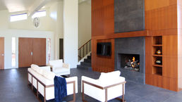 Modern living room interior in a remodeled Sedona home featuring a contemporary fireplace with a dark porcelain tile surround, tall ribbon mahogany wood panels, a modern log set, and white lounge seating.