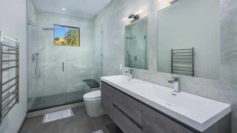 Modern guest bathroom in a custom Sedona home featuring a dual-sink vanity with wood-grain cabinets, a large glass-enclosed walk-in shower, light gray wall tiles, and a metal heated towel rack.