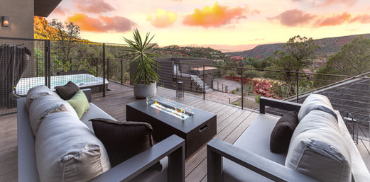 Modern outdoor deck of a custom home in Sedona during sunset, featuring plush seating, a fire pit table, and sweeping views of the red rock landscape under a golden sky.