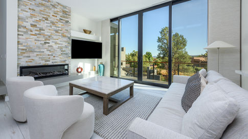 Modern living room in a custom Sedona home featuring a textured stone feature wall with an integrated TV and linear fireplace, white armchair seating, a light gray rug, and expansive glass doors showcasing natural views.
