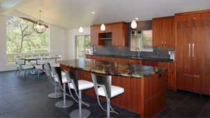 Bright, remodeled open-concept kitchen in a custom Sedona home featuring warm ribbon mahogany cabinets, a large granite island with white barstools, contemporary pendant lighting, and a dining area with scenic window views.