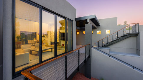 Modern custom Sedona home exterior at twilight viewed from an elevated deck, featuring large glass doors glowing with warm interior light, stucco walls, and sleek metal railings against a sunset sky.