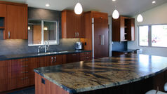 Close-up view of a remodeled kitchen island featuring a dramatic high-movement brown and cream granite countertop, warm ribbon mahogany cabinets, a stainless steel sink, and modern pendant lighting.