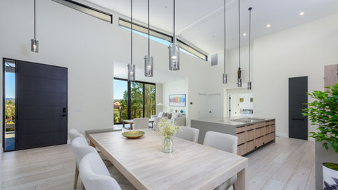 Modern open-concept dining area in a custom Sedona home featuring a light wood dining table with white chairs, high ceilings with trapezoid windows, minimalist pendant lights, and a view into the adjacent kitchen.