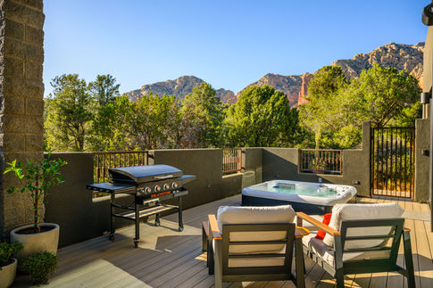 Sunny daytime view of a Southwest Contemporary Sedona outdoor deck remodel featuring a built-in grill, comfortable lounge seating, and a relaxing spa against a beautiful red rock mountain backdrop.