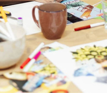 A high-angle, close-up view of a wooden table covered in art supplies, coffee mugs, and colorful collages, with participants gathered around to collaborate during a workshop.