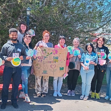 A group of diverse adults standing in a row holding their watercolor paintings and a community mural on bown paper. Behind them is trees and a bright blue sky.